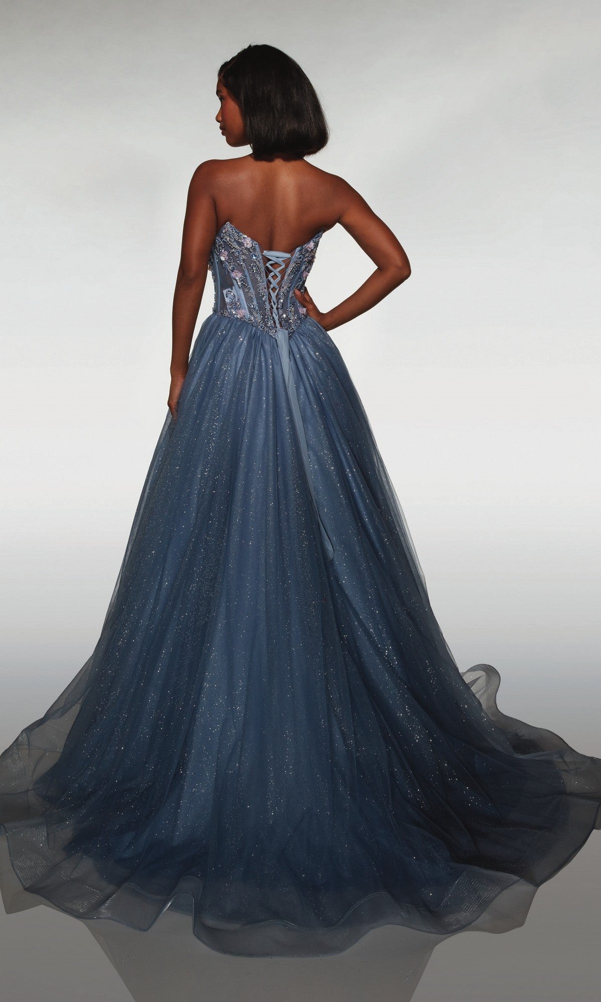 A woman models the Alyce 62009, a strapless, floor-length blue glitter tulle prom gown with a lace-up corset bodice and full skirt, standing with her back to the camera.